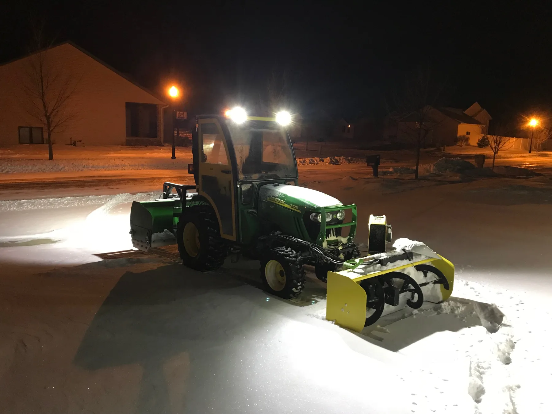 A Baker Snow & Ice tractor clearing a residential driveway at night with work lights on